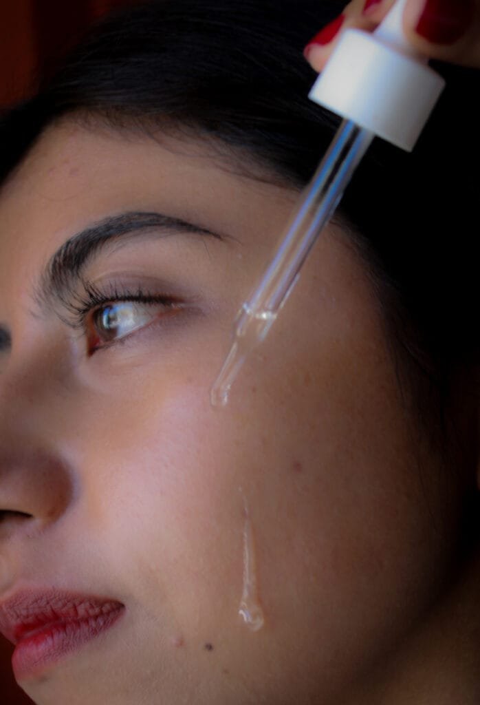 Mujer joven de perfil aplicando sérum facial en la mejilla con un gotero.